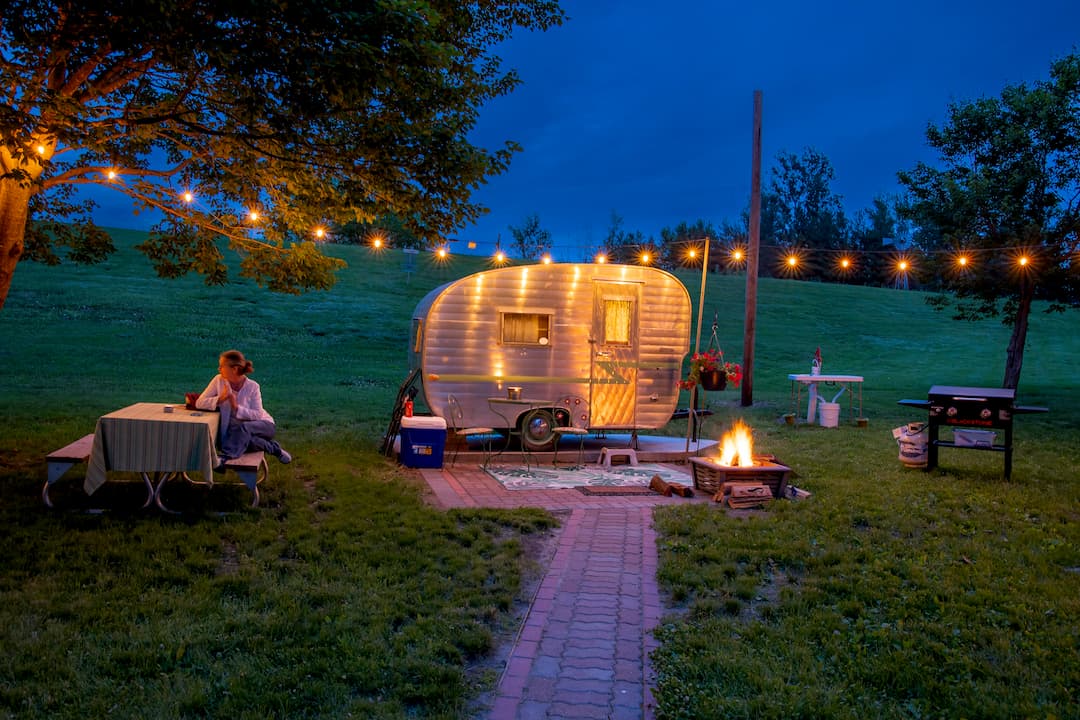 Vintage camper glowing under string lights beside a campfire at dusk
